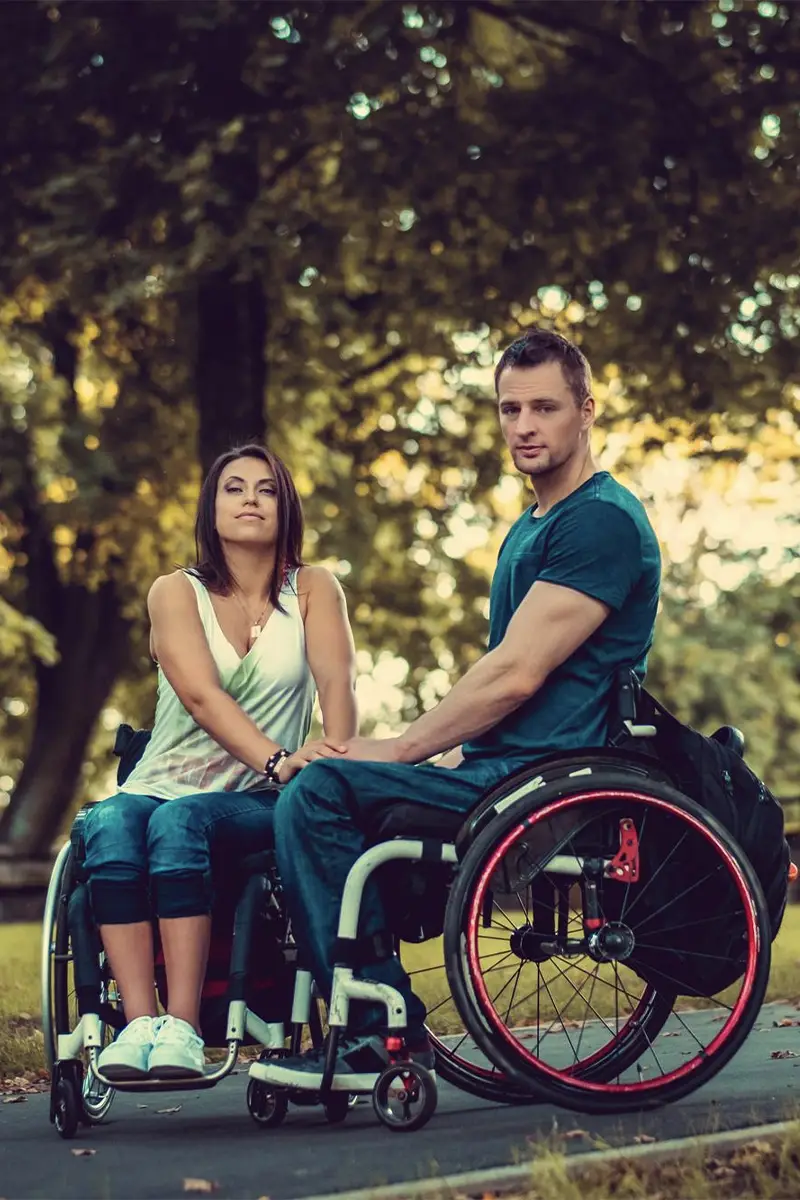 Dwie osoby siedzące na wózkach trzymają się za ręce. W tle widać zielone drzewa