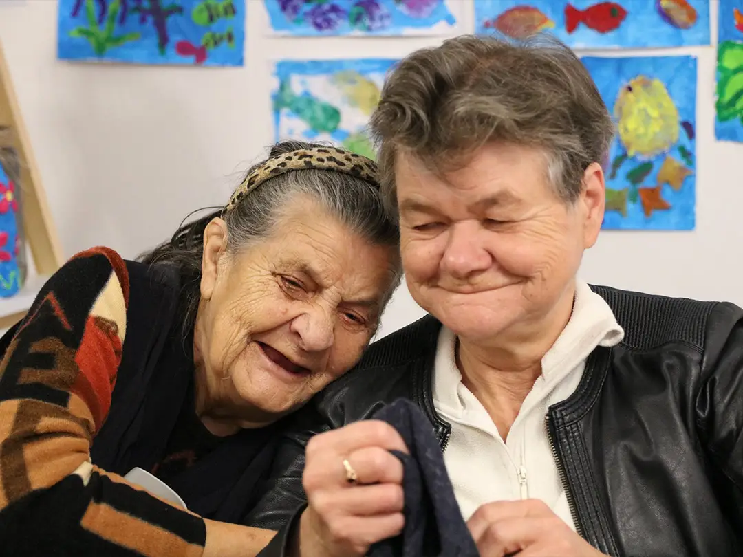 Dwie starsze panie, jedna ubrana w kolorowy sweter, druga w skórzaną kurtkę siedzą uśmiechnięte i przytulone na tle rysunków zawieszonych na ścianie. Osoby patrzą na kawałek materiału trzymany przez jedną z nich