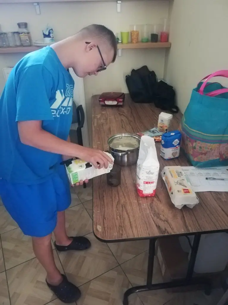 Chłopiec w niebieskim T-shircie i krótkich spodenkach stoi przy stole i oburącz wlewa mleko do szklanki. Na stole stoją składniki na ciasto oraz garnek z masą