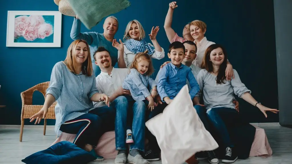 Siedząca na kanapie grupa osób, ubrana w białe, niebieskie i błękitne ubrania i podrzucająca w powietrze poduszki. W tle widać granatową ścianę, krzesło i obrazek na ścianie